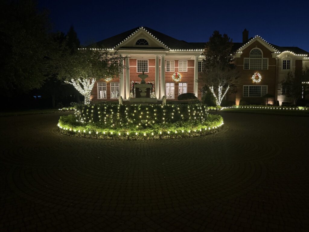 Brick house with warm white roofline lights and wreaths behind a circular hedge wrapped in lights