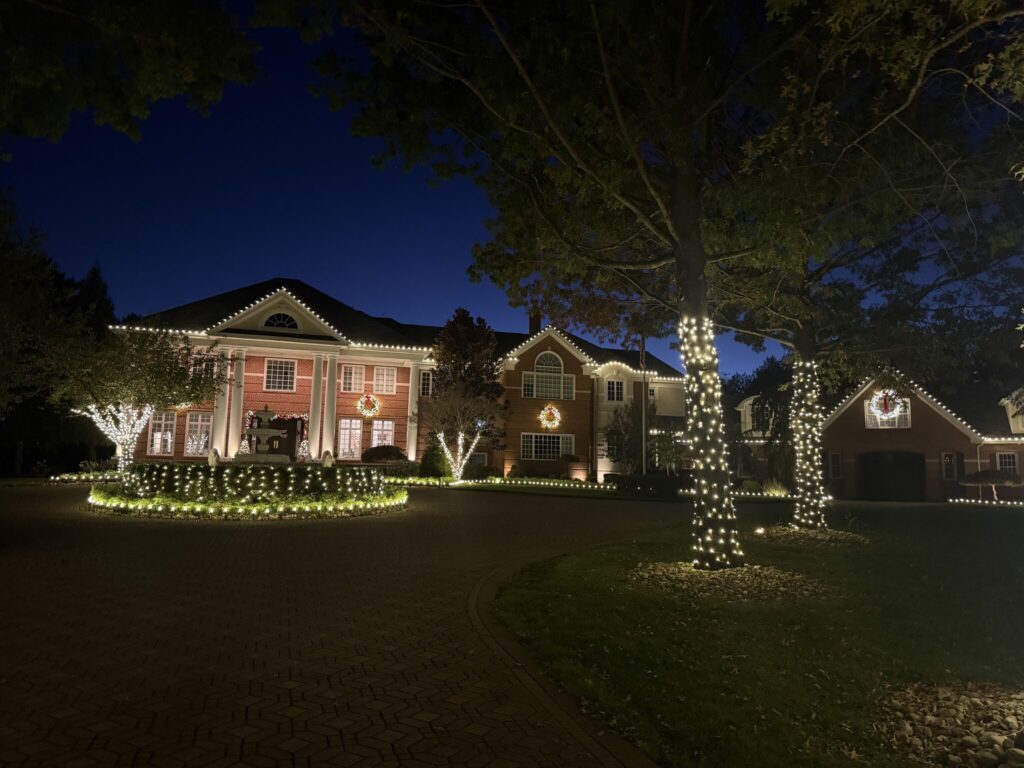 Brick estate at night with warm white roofline lights, wreaths, wrapped trees and a lit hedge circle