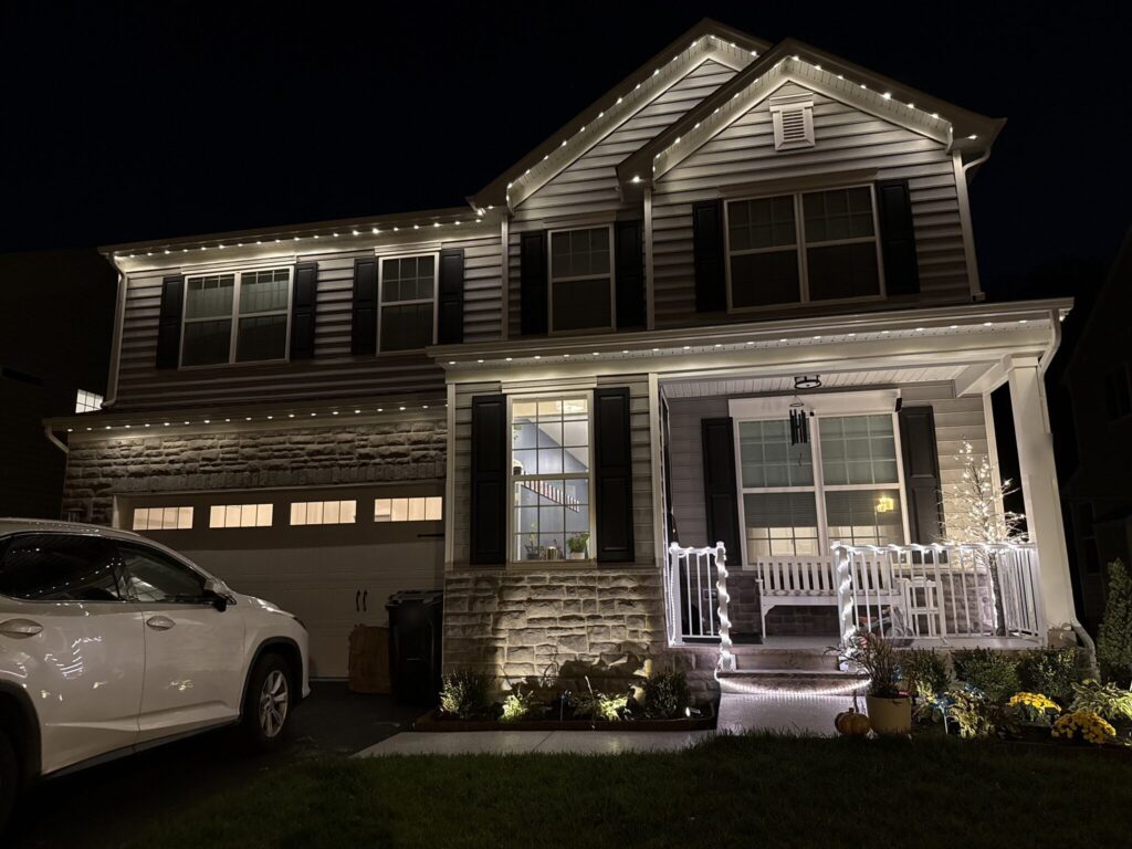 Two-story house at night with warm white permanent lights along every roofline and a lit porch railing